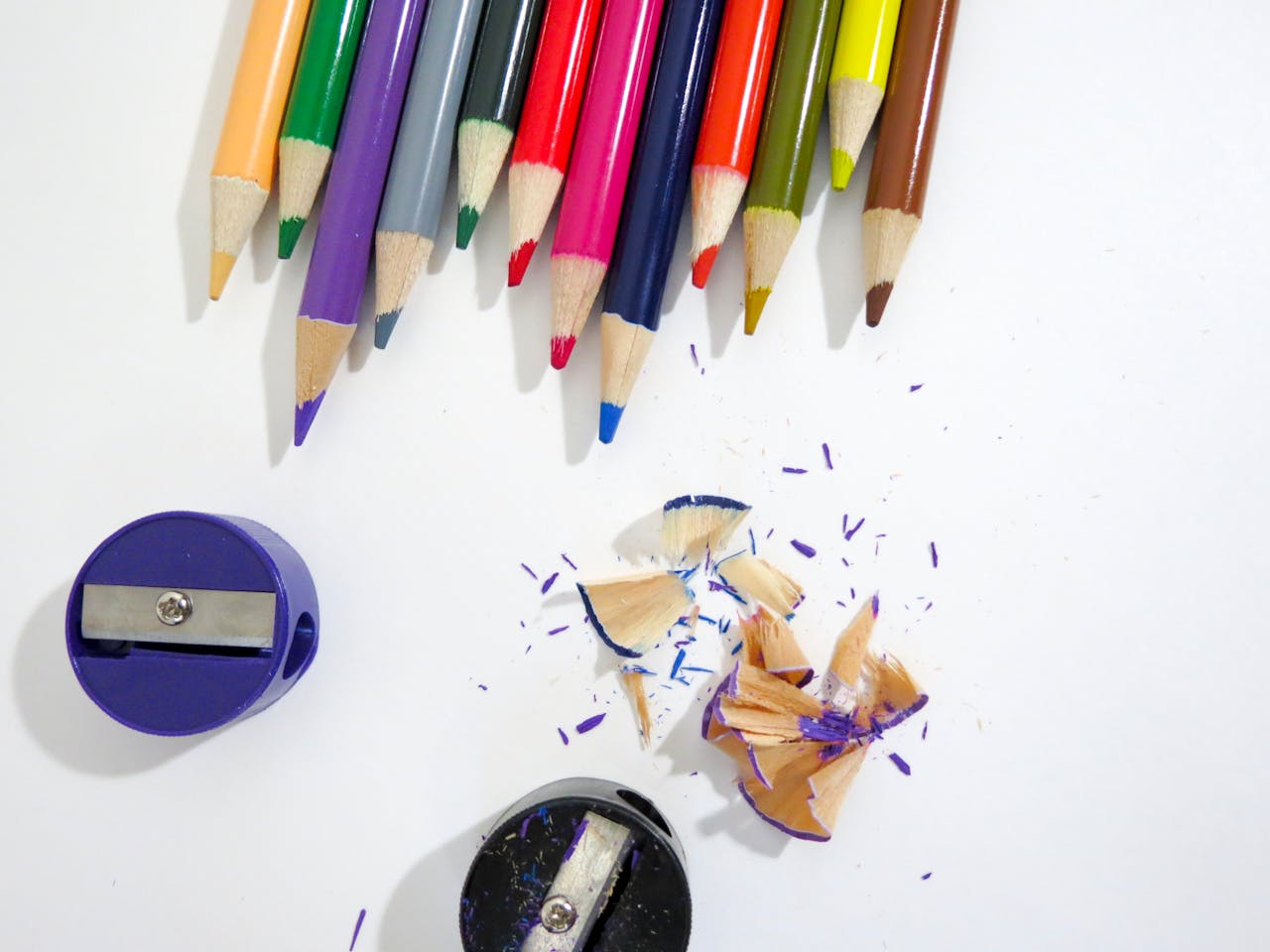 Bright colored pencils and sharpeners with shavings on a white surface.